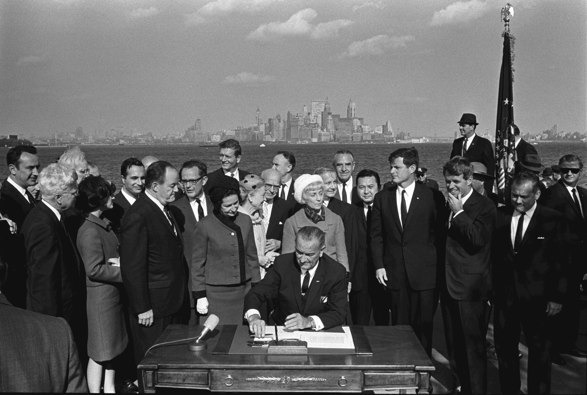 President Lyndon B. Johnson signs the Immigration and Nationality Act as Vice President Hubert Humphrey, Lady Bird Johnson, Muriel Humphrey, Sen. Edward (Ted) Kennedy, Sen. Robert F. Kennedy, and others look on.